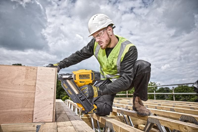 A construction worker wearing a safety helmet and high-visibility vest is using a yellow DEWALT DCN930N nailer to secure wooden boards at an outdoor building site. The worker's face is blurred for privacy.