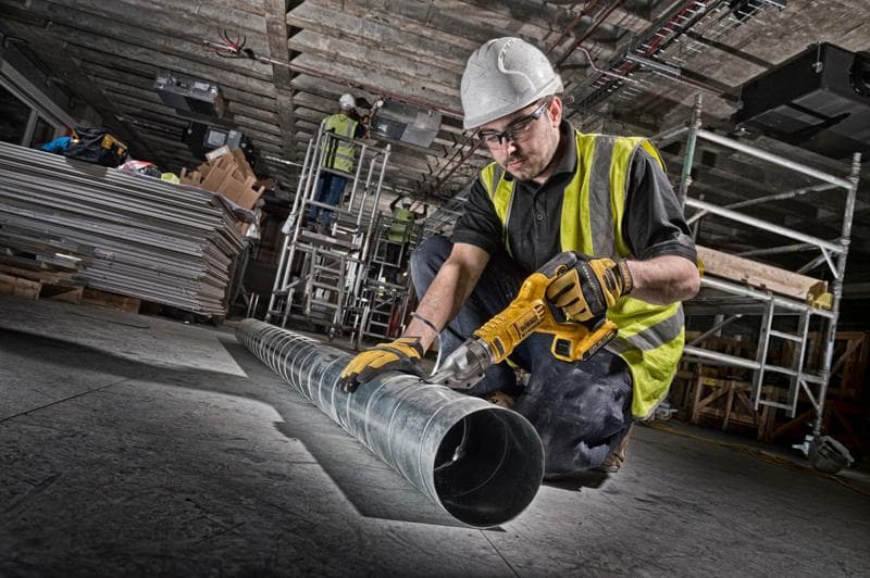 A worker wearing a yellow safety vest and white helmet is using a DEWALT cordless metal shear (model DCS491N) to cut a long metal duct in an industrial construction environment.