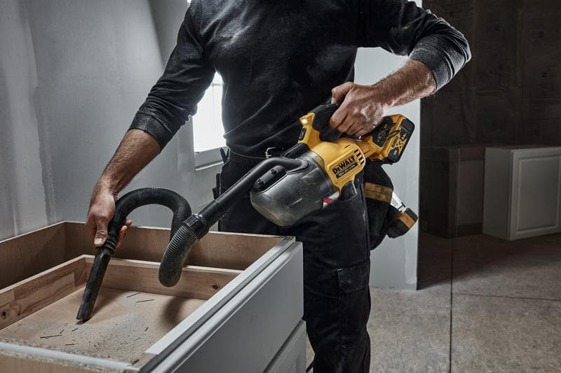 A person using a DEWALT cordless handheld vacuum cleaner (model DCV501LN) to clean sawdust from the surface of a wooden cabinet in a workshop or renovation setting.
