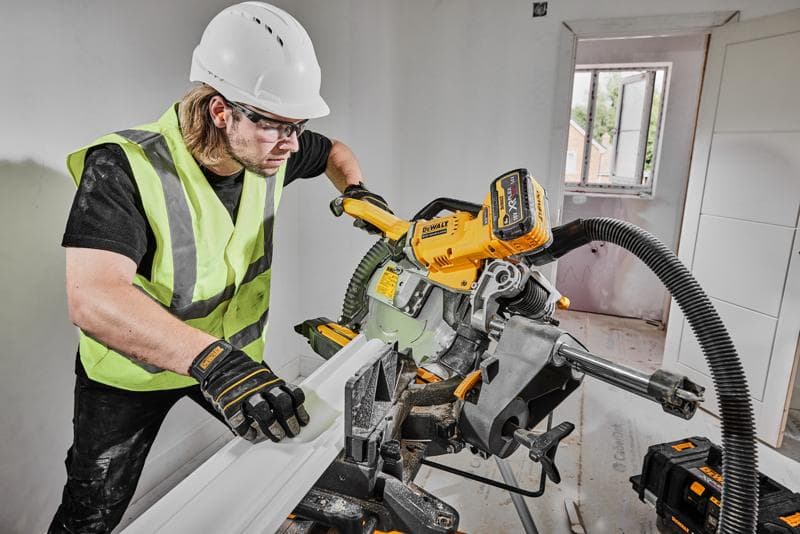 A person wearing a safety vest, gloves, and hard hat is using a DEWALT DCS781X2 cordless miter saw to cut a piece of trim inside a room under construction. The saw is connected to a dust extraction system, and there are DEWALT tool batteries visible nearby.