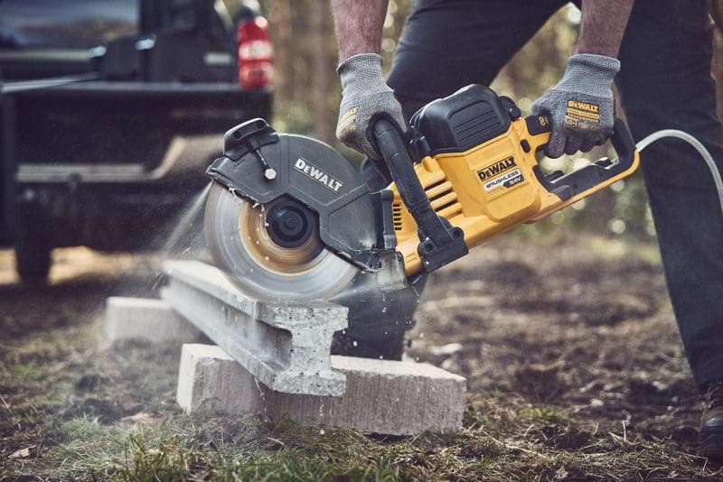 A person wearing gloves uses a DEWALT cordless circular saw to cut a concrete block outdoors. The saw is generating dust as