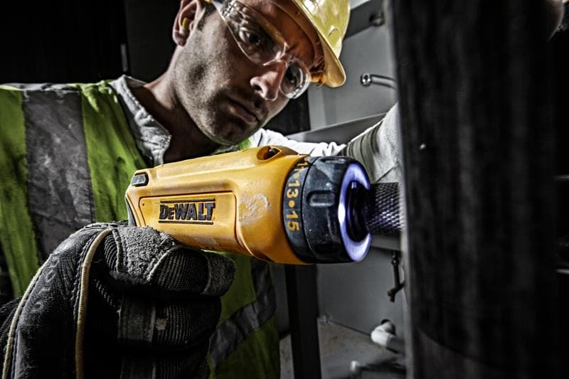 A person wearing gloves and a safety vest uses a yellow DEWALT electric screwdriver (model DCF680G2) with an illuminated tip, working on a piece of equipment in an industrial setting.