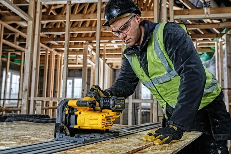 A worker using a DEWALT DCS520T2-XE cordless track saw in a construction site with wooden framing in the background. The worker is wearing a safety vest, gloves, and a hard hat.