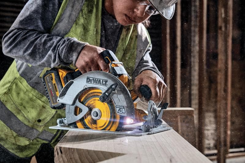 A person wearing a safety vest and helmet is using a DEWALT DCS573NT cordless circular saw to cut through a wooden plank in a construction setting.