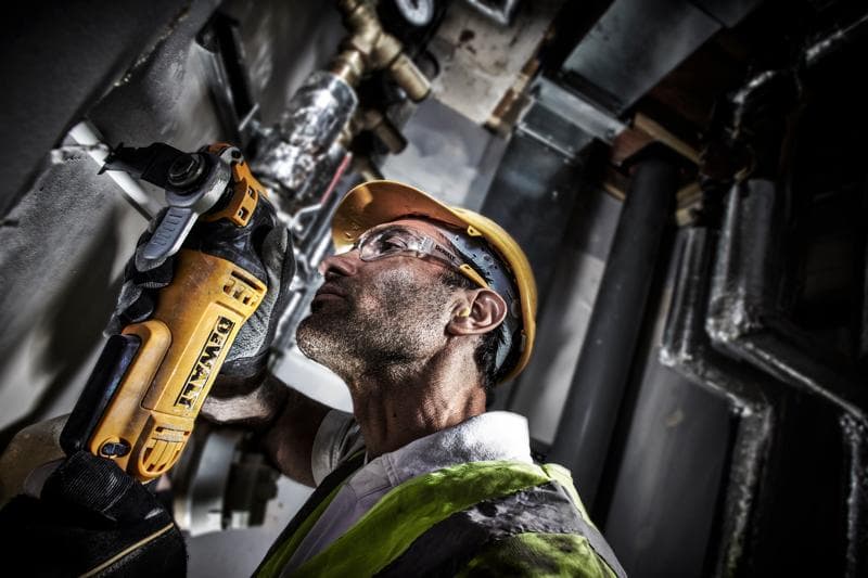 A person wearing a yellow hard hat and safety vest uses a DEWALT DWE315 power tool to cut or grind material on a wall in an industrial setting. Their face is obscured.
