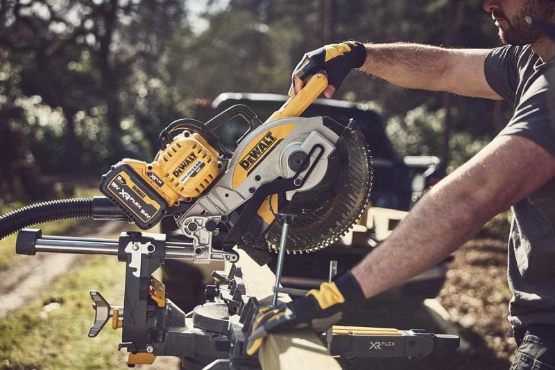 A person using a DEWALT DCS727N cordless mitre saw outdoors to cut a piece of wood, wearing protective gloves. The yellow