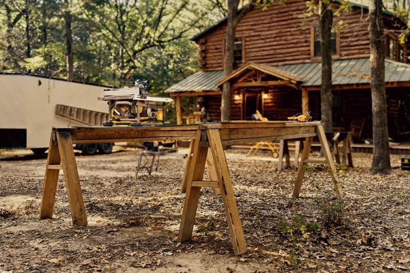 A DEWALT DWS780 miter saw placed on a wooden workbench outdoors, with a rustic log cabin in the background surrounded by trees and autumn leaves.