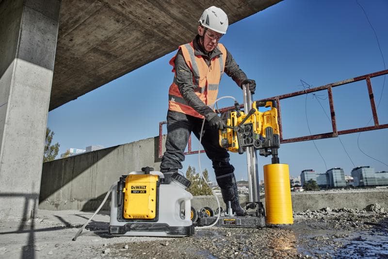 A person wearing protective gear and an orange safety vest is operating a DEWALT concrete drilling machine outdoors at a construction site. The DEWALT equipment, including a yellow drill and a white water tank with the DEWALT logo, is positioned on a concrete surface under an unfinished structure.