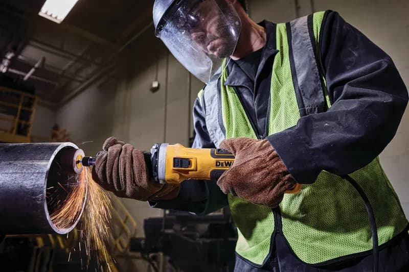 Worker using a DEWALT angle grinder to cut a metal pipe, sparks flying, wearing gloves and safety gear indoors.