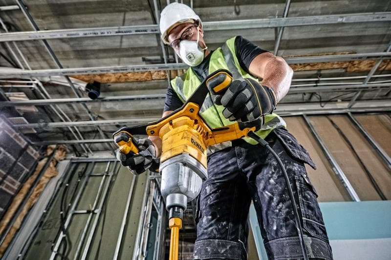 A construction worker wearing a safety vest, gloves, and a hard hat uses a yellow DEWALT corded mixing drill (DWD241) inside a building under construction.