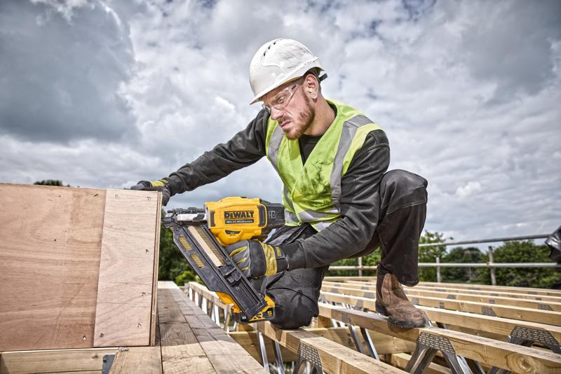 A construction worker wearing a safety helmet and high-visibility vest uses a DEWALT DCN930N cordless nailer to attach wooden boards at a building site. The worker is kneeling on wooden beams with a cloudy sky in the background.
