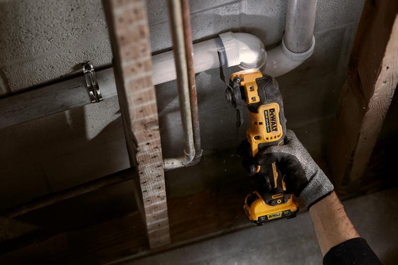 A person wearing a glove uses a DEWALT cordless oscillating multi-tool to cut a white PVC pipe near a wall with exposed pipes and beams.