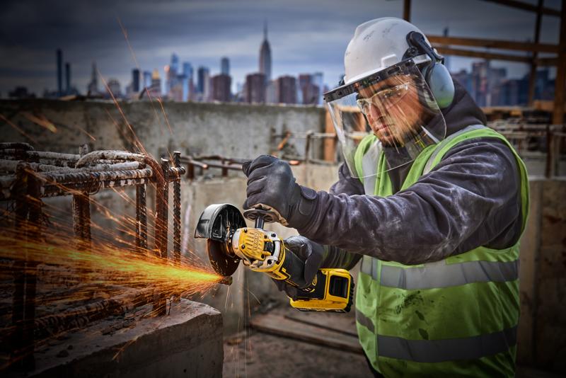 A construction worker wearing safety gear uses a DEWALT DCG405P2 cordless angle grinder to cut metal rebar, with sparks flying, at an outdoor construction site in the city.