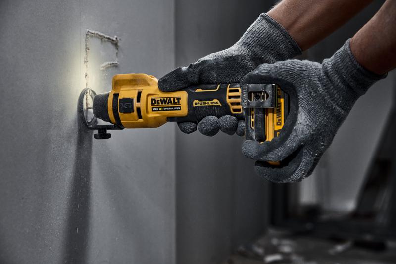 A person wearing grey gloves is using a DEWALT cordless rotary tool to cut a square hole in a drywall panel.