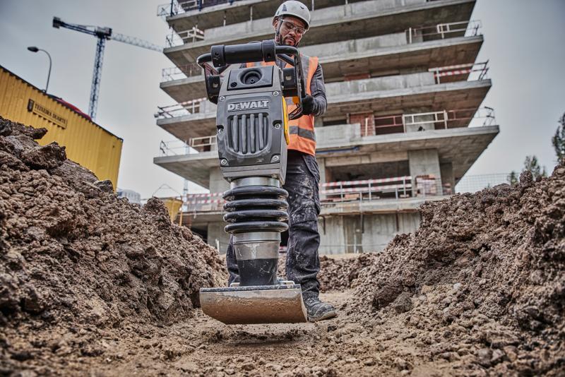 A DEWALT compactor machine being operated by a construction worker wearing a safety helmet and orange vest, working on a dirt trench at a construction site with a partially built structure and equipment in the background.