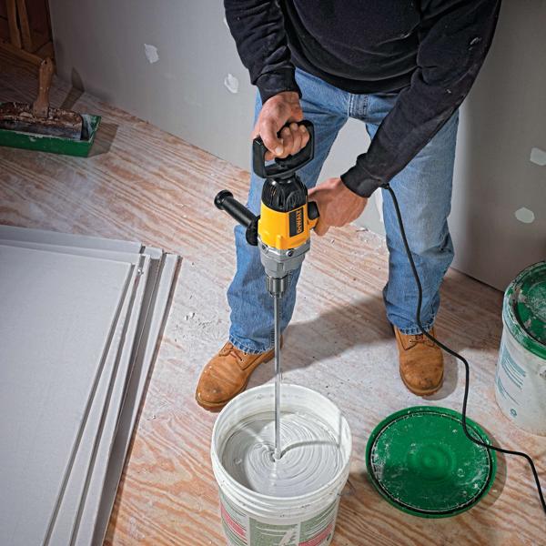 A person using a DEWALT electric drill mixer to mix white compound in a large bucket on a construction site. The person is standing on a wooden floor, wearing work boots, jeans, and a black sweatshirt. There are sheets of drywall and construction materials nearby.