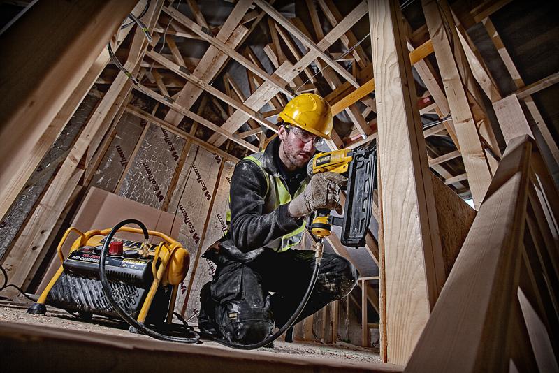 A person wearing a yellow hard hat and safety vest is working on a wooden construction site, using a DEWALT pneumatic nail gun connected to a DEWALT air compressor. The person is crouched among wooden beams, and their face is blurred.