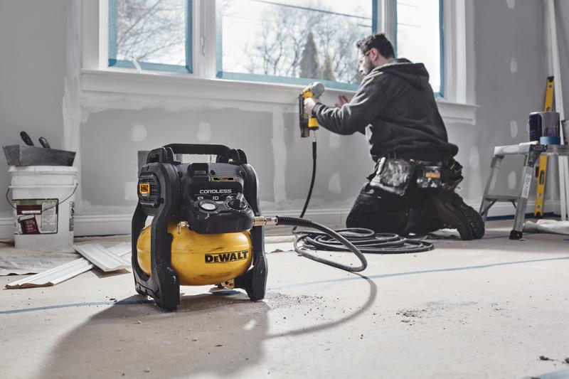 A DEWALT cordless air compressor is placed on the floor in a room under renovation. In the background, a person is kneeling